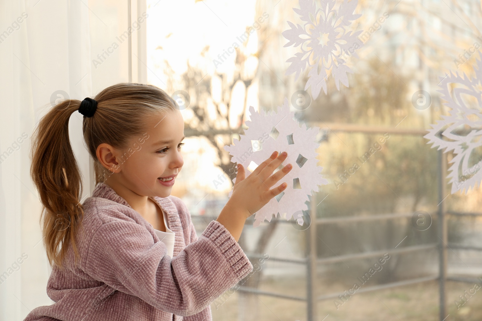 Photo of Little girl decorating window with paper snowflake indoors