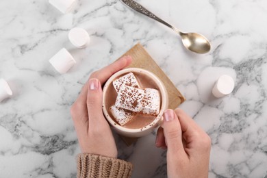 Photo of Woman drinking aromatic hot chocolate with marshmallows and cocoa powder at white marble table, top view
