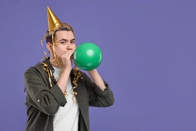 Young man with party hat and balloon on purple background, space for text