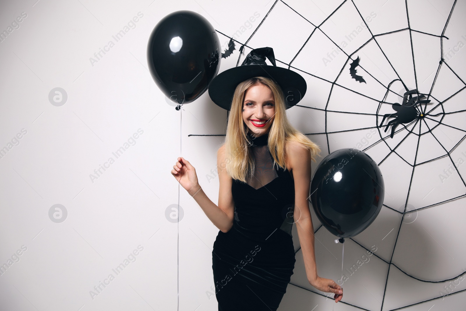 Photo of Woman in witch hat with balloons posing near white wall decorated for Halloween