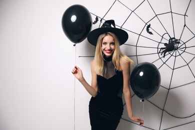 Photo of Woman in witch hat with balloons posing near white wall decorated for Halloween