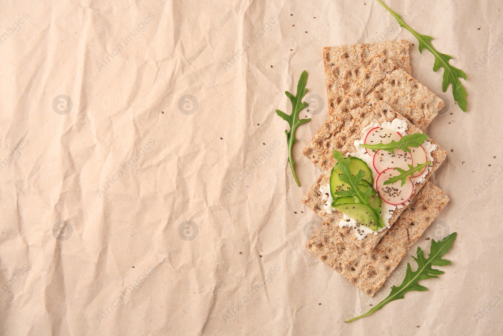 Photo of Tasty crispbreads with cream cheese, fresh cucumbers, radish and arugula on parchment paper, flat lay. Space for text