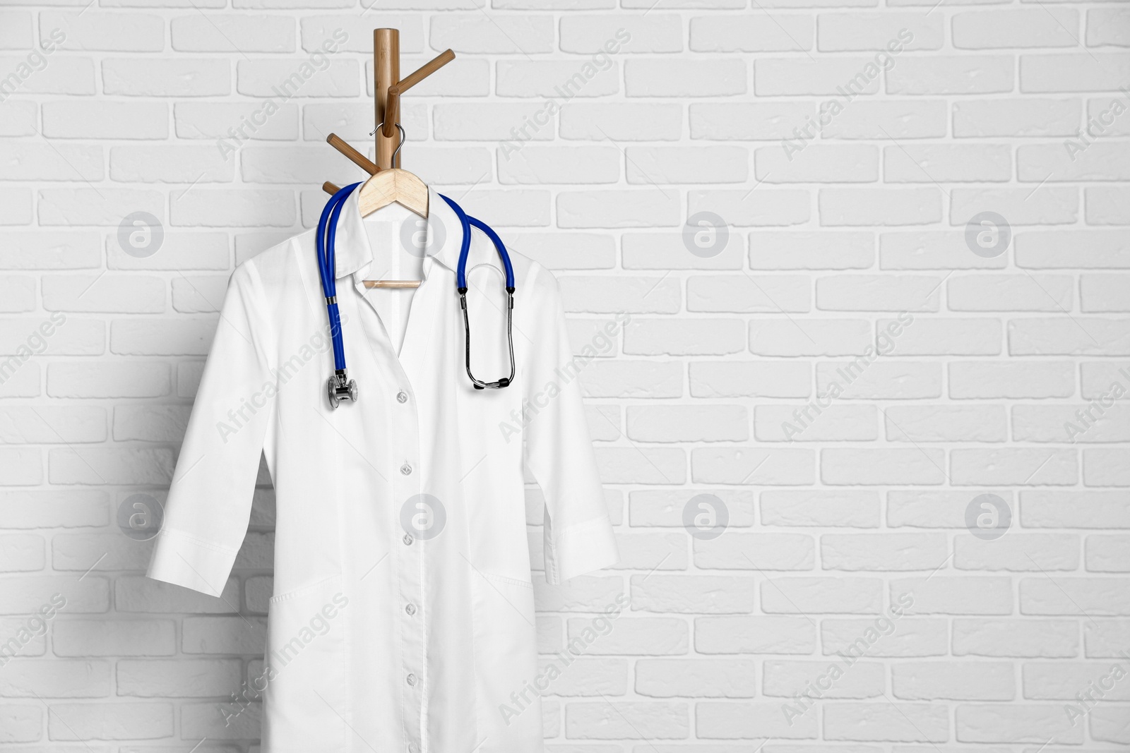 Photo of Medical uniform and stethoscope hanging on rack near white brick wall. Space for text