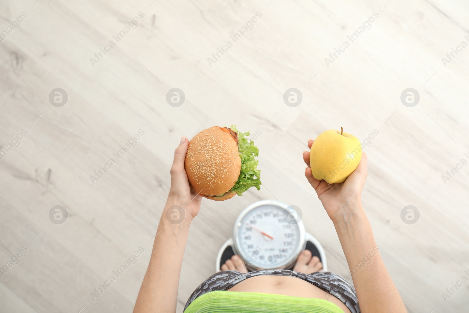 Photo of Woman holding tasty sandwich and fresh apple while measuring her weight on floor scales, top view. Choice between diet and unhealthy food