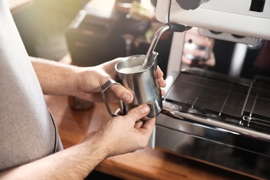 Photo of Barista frothing milk in metal pitcher with coffee machine wand at bar counter, closeup