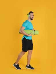 Handsome man with yoga mat and headphones on yellow background