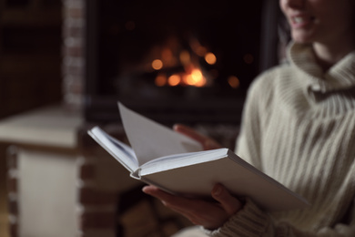 Photo of Woman reading book near burning fireplace at home, closeup