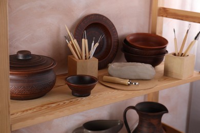 Set of different crafting tools and clay dishes on wooden rack in workshop