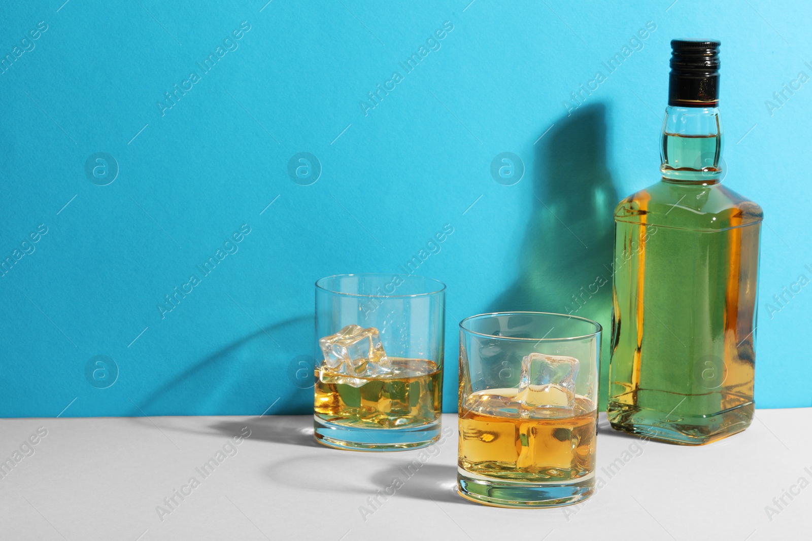 Photo of Whiskey with ice cubes in glasses and bottle on white table against light blue background, space for text