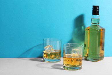 Photo of Whiskey with ice cubes in glasses and bottle on white table against light blue background, space for text