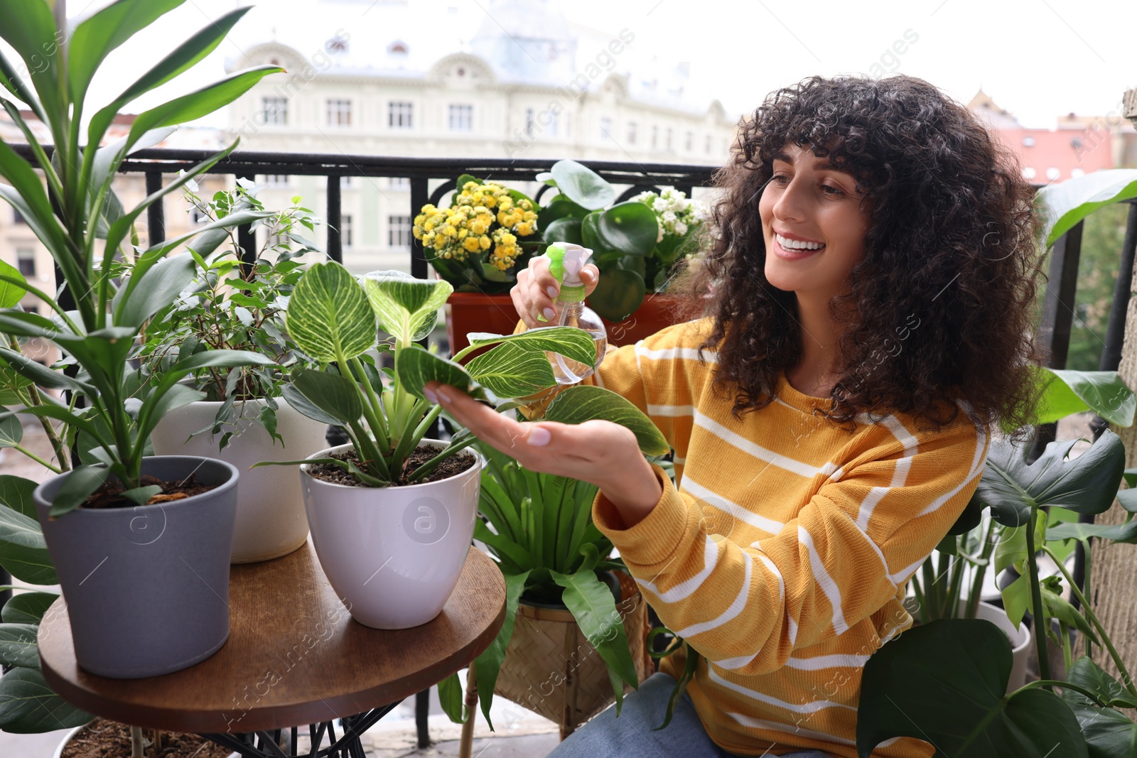 Photo of Beautiful young woman spraying potted houseplants with water on balcony