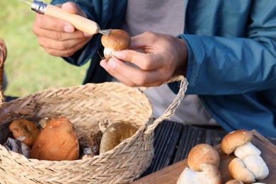 Photo of Man cleaning mushroom with brush on knife at table outdoors, closeup