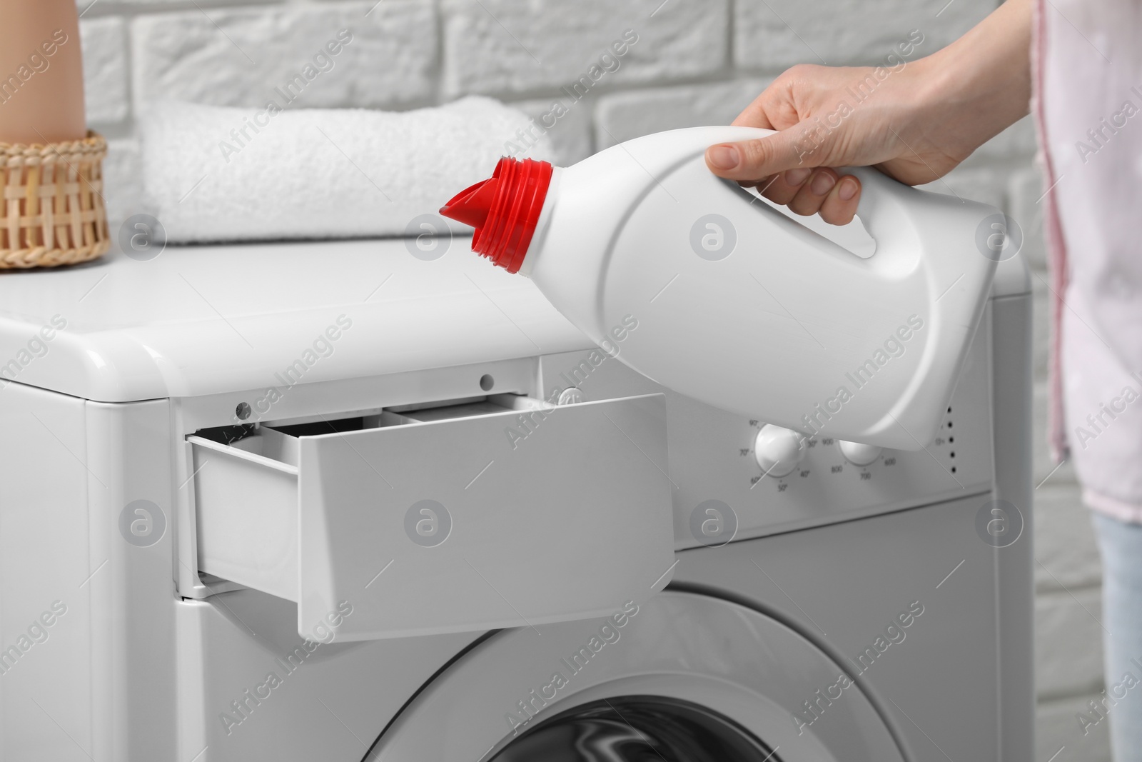 Photo of Woman pouring fabric softener from bottle into washing machine near white brick wall, closeup