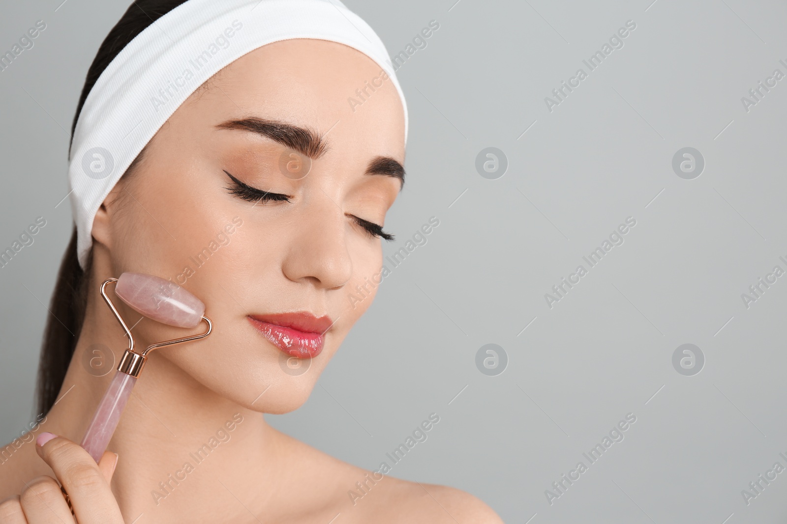 Photo of Woman using natural pink quartz face roller on grey background, closeup. Space for text