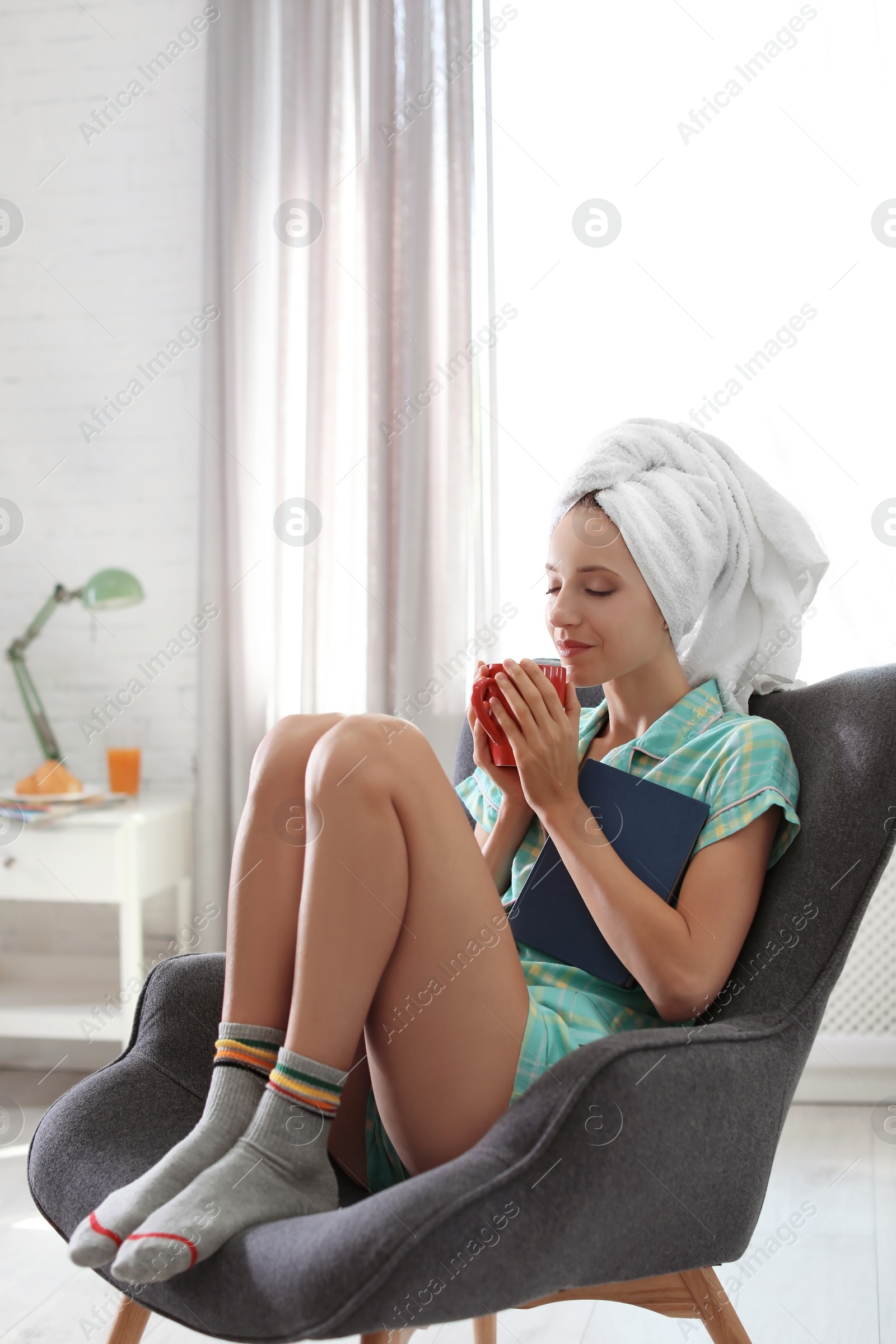Photo of Young happy woman with book and cup of aromatic coffee in armchair at home. Lazy morning