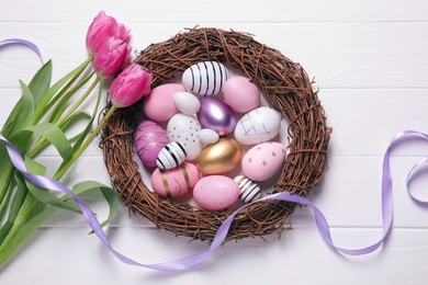 Many painted Easter eggs, tulip flowers and ribbon on white wooden table, flat lay