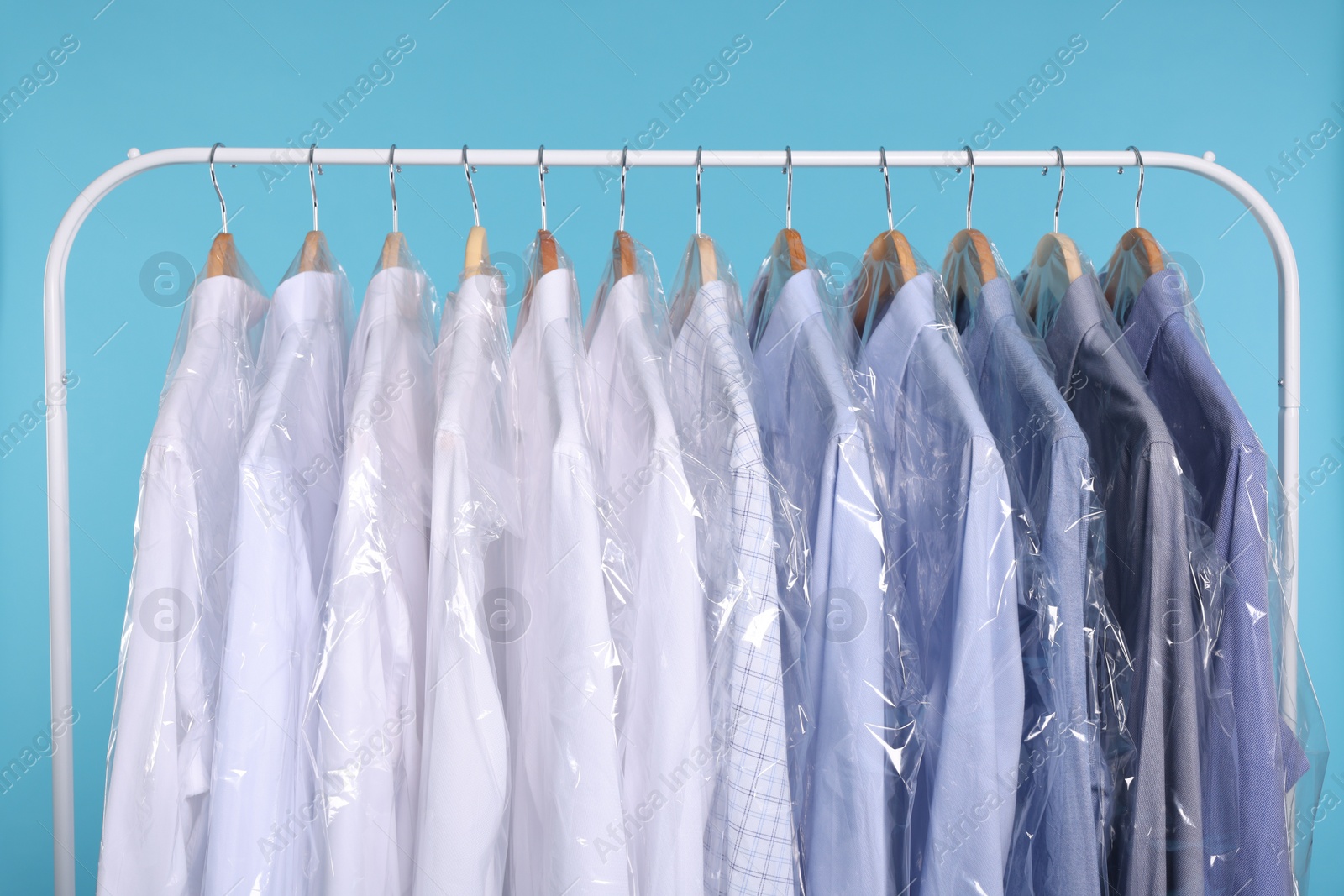 Photo of Dry-cleaning service. Many different clothes in plastic bags hanging on rack against light blue background