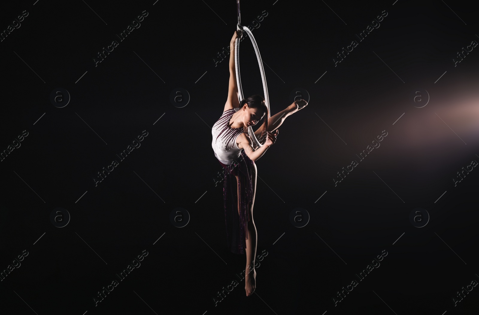 Photo of Young woman performing acrobatic element on aerial ring against dark background