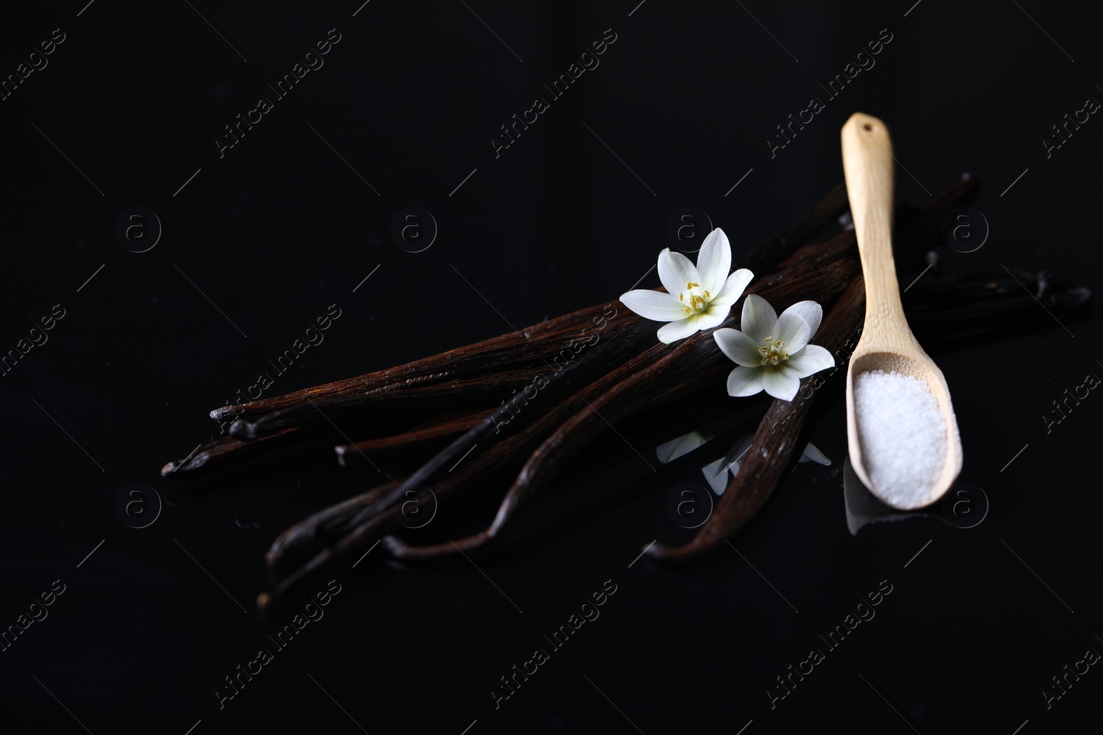 Photo of Vanilla pods, flowers and spoon with sugar on dark mirror table, closeup