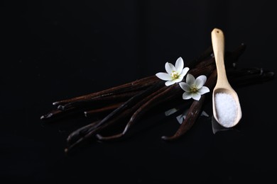 Photo of Vanilla pods, flowers and spoon with sugar on dark mirror table, closeup