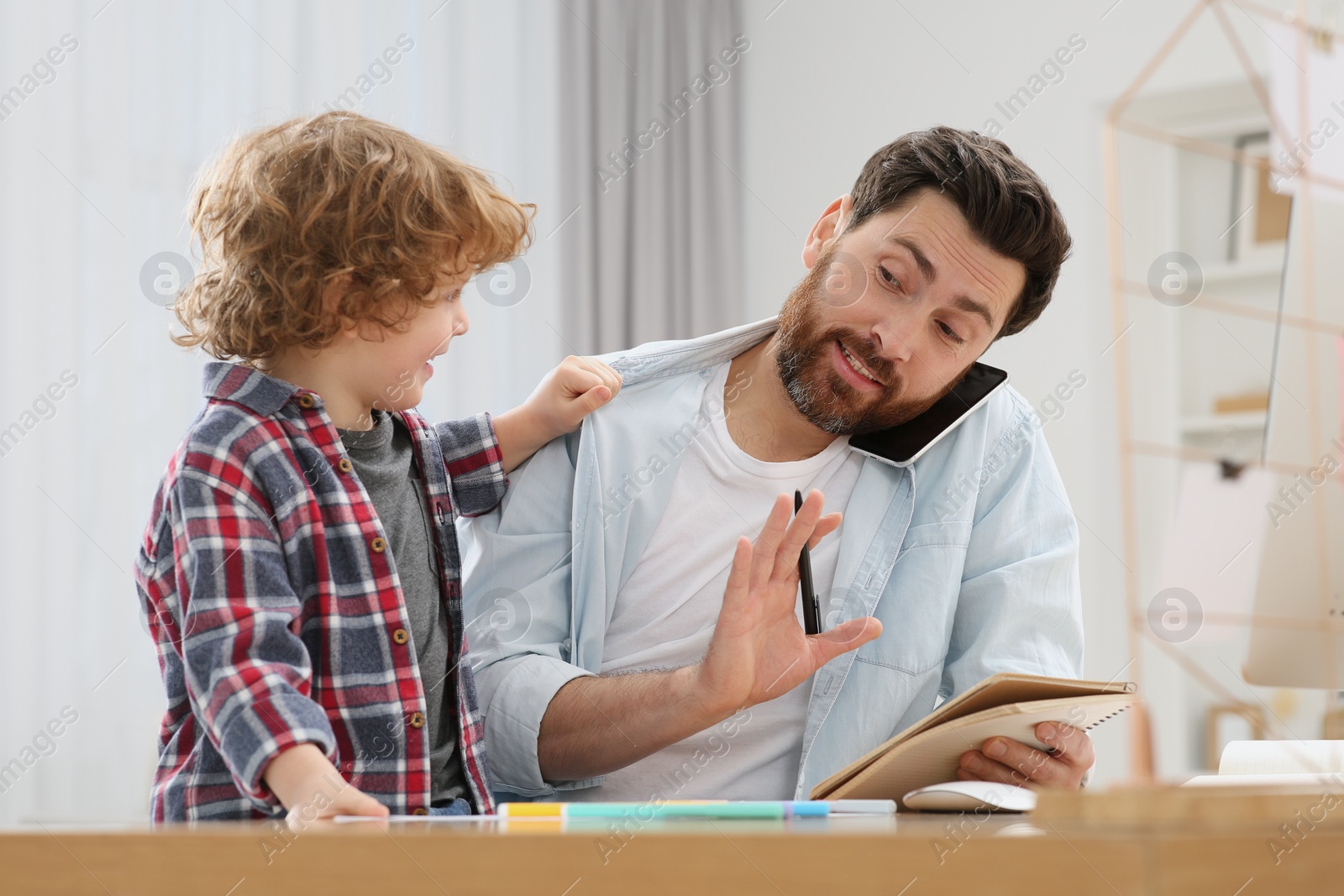 Photo of Little boy bothering his father at home. Man working remotely at desk