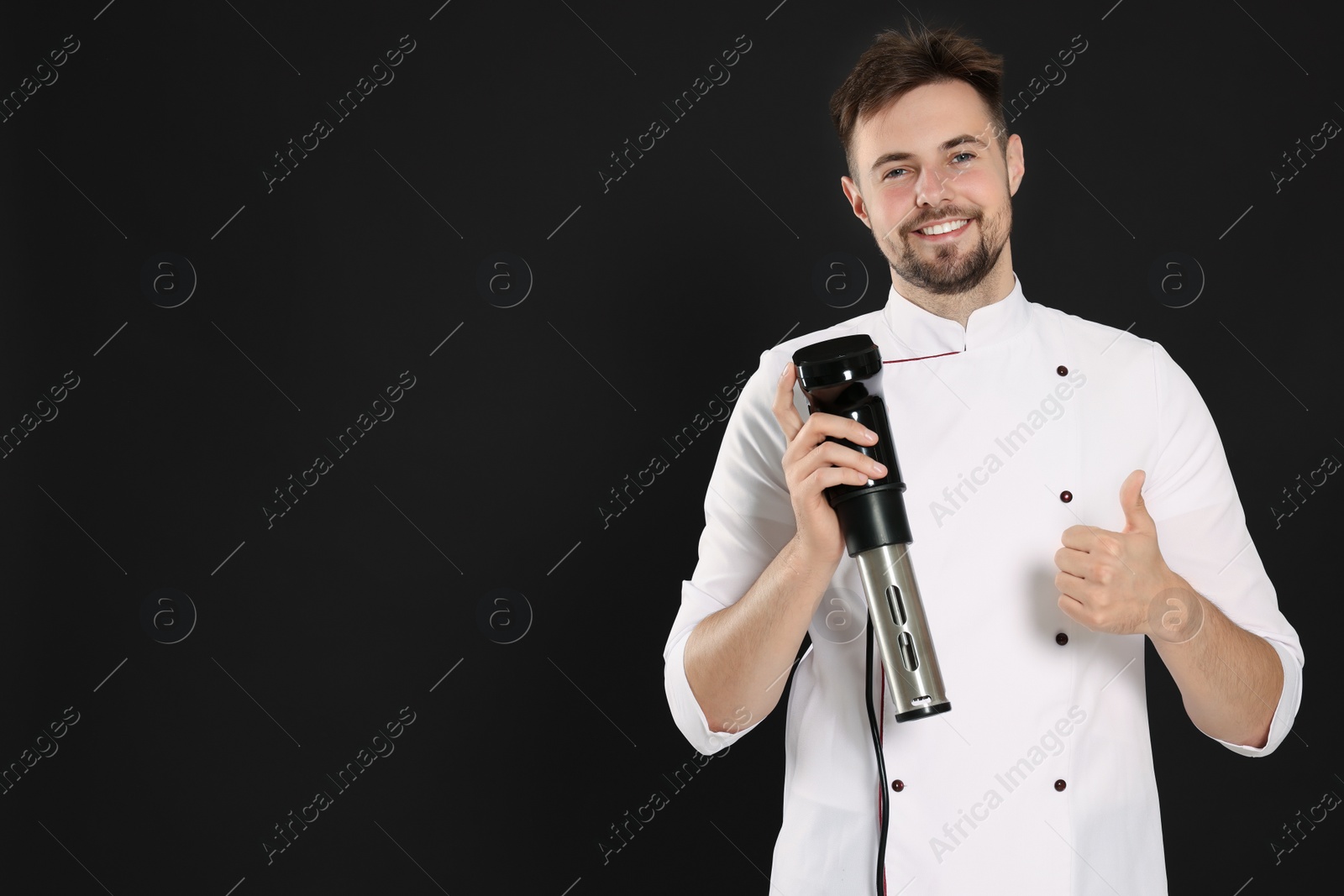 Photo of Smiling chef holding sous vide cooker and showing thumb up on black background, space for text