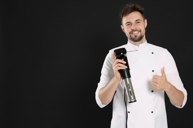 Smiling chef holding sous vide cooker and showing thumb up on black background, space for text