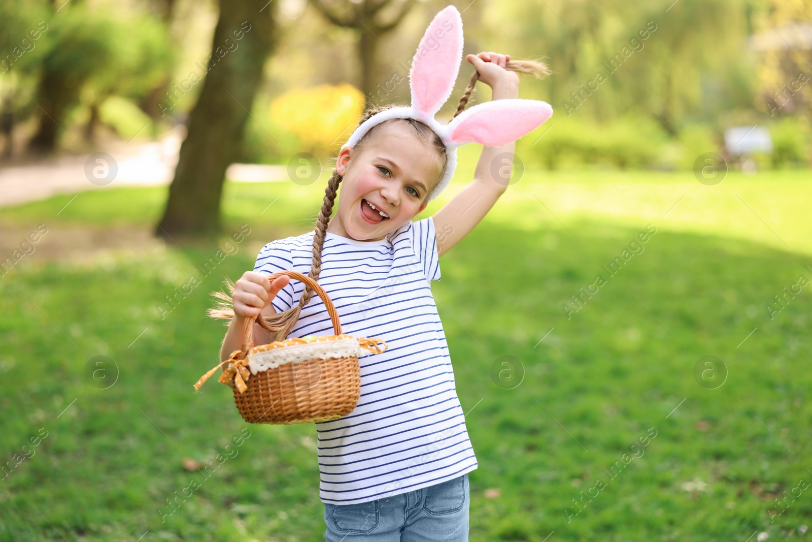 Photo of Easter celebration. Cute little girl in bunny ears holding wicker basket outdoors