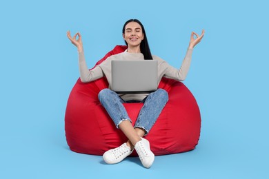 Happy woman with laptop sitting on beanbag chair and meditating against light blue background