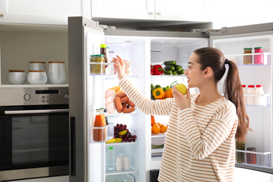 Photo of Young woman with apple near open refrigerator in kitchen