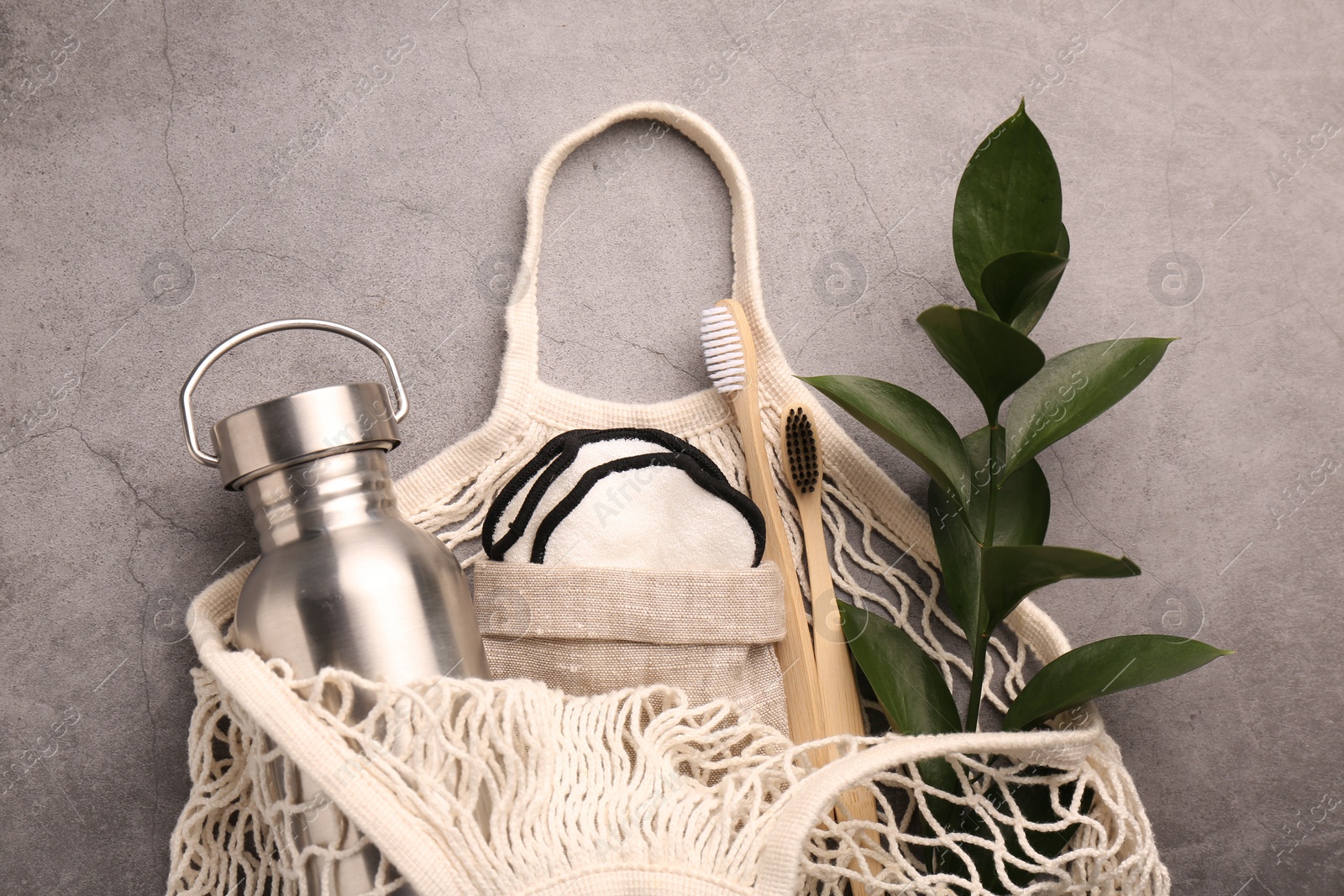 Photo of Fishnet bag with different items and green twig on grey table, top view. Conscious consumption