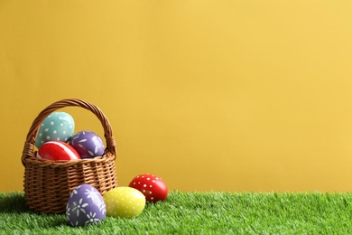 Photo of Wicker basket with painted Easter eggs on green grass against color background, space for text