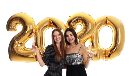 Happy young women with glasses of champagne near golden 2020 balloons on white background. New Year celebration