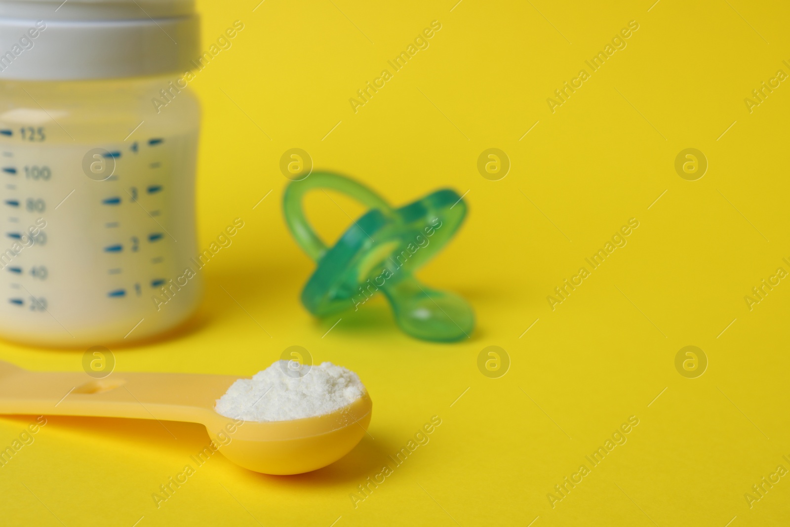 Photo of Scoop of powdered infant formula, feeding bottle and pacifier on yellow background, space for text. Baby milk