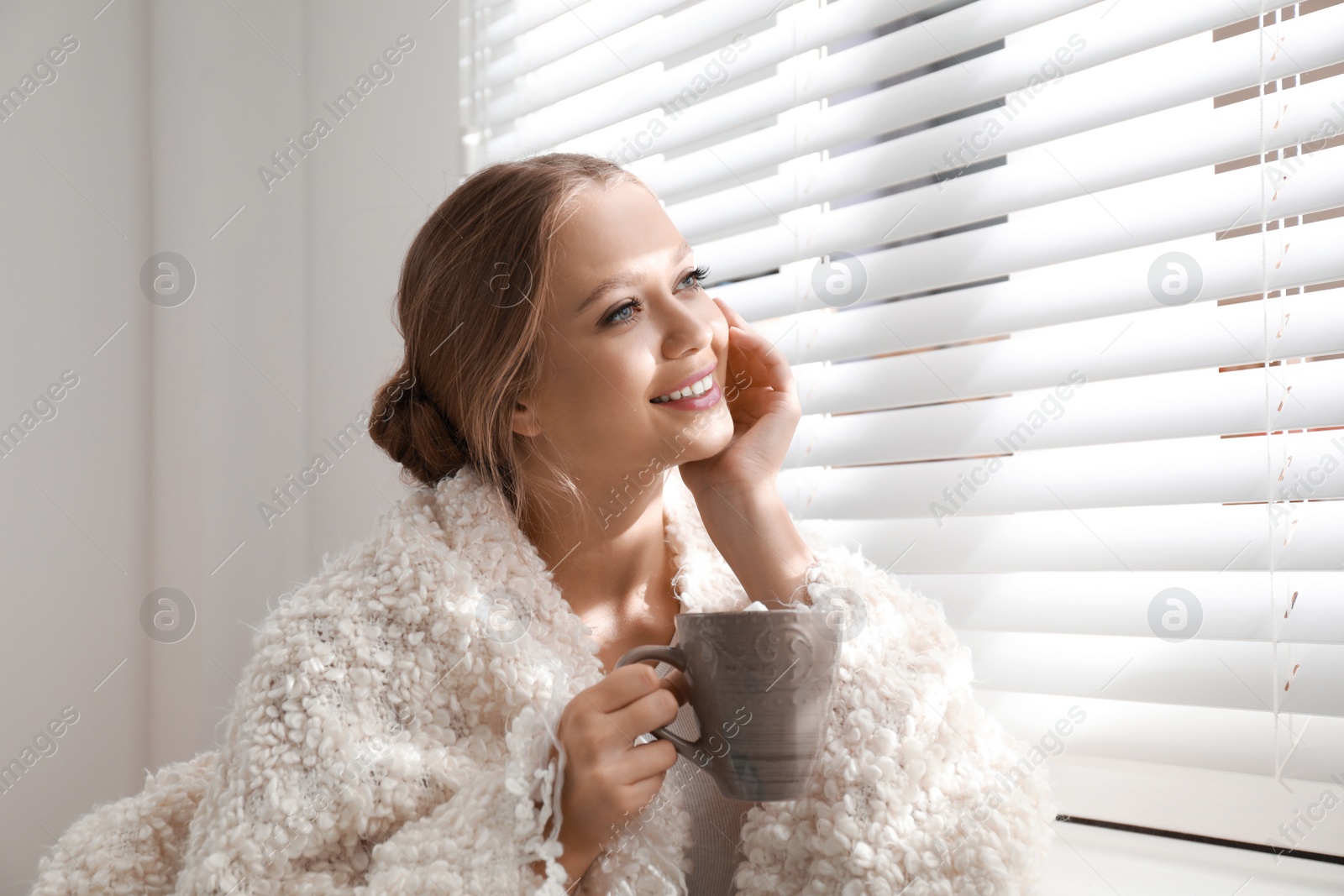 Photo of Beautiful young woman with cup of hot drink near window at home. Winter atmosphere