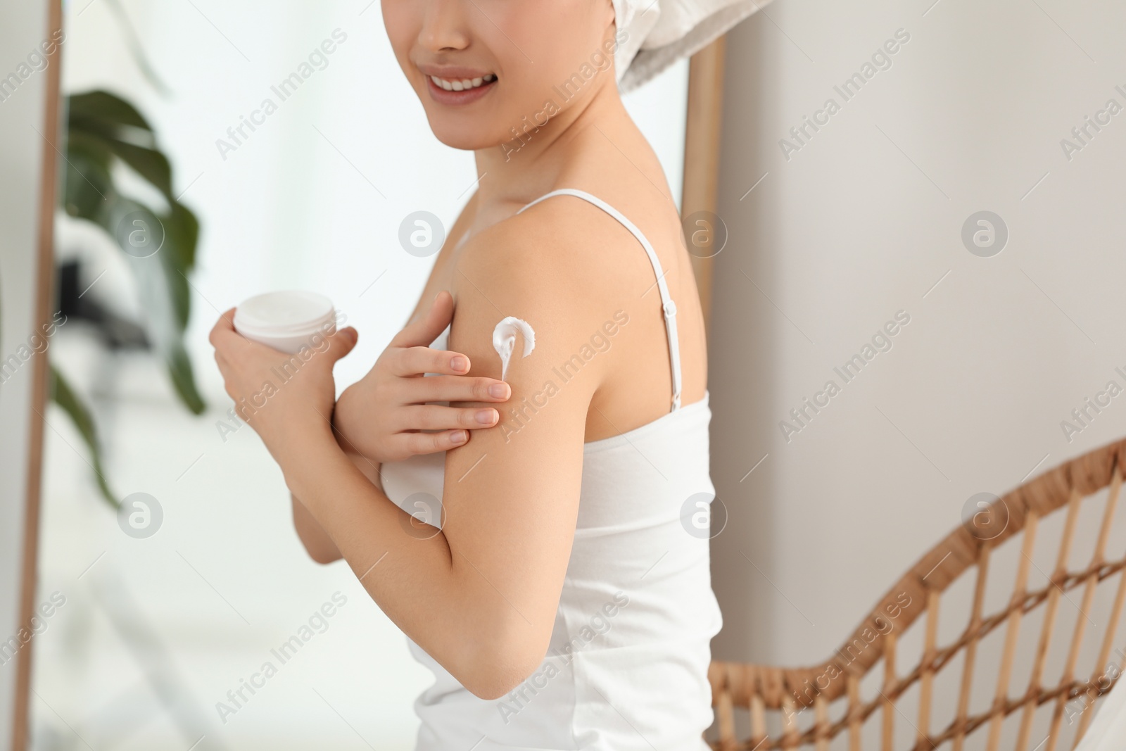 Photo of Young woman applying body cream on shoulder in room, closeup