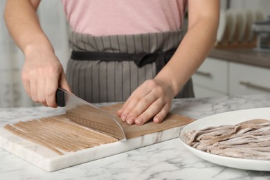 Photo of Woman making soba (buckwheat noodles) with knife at white marble table in kitchen, closeup