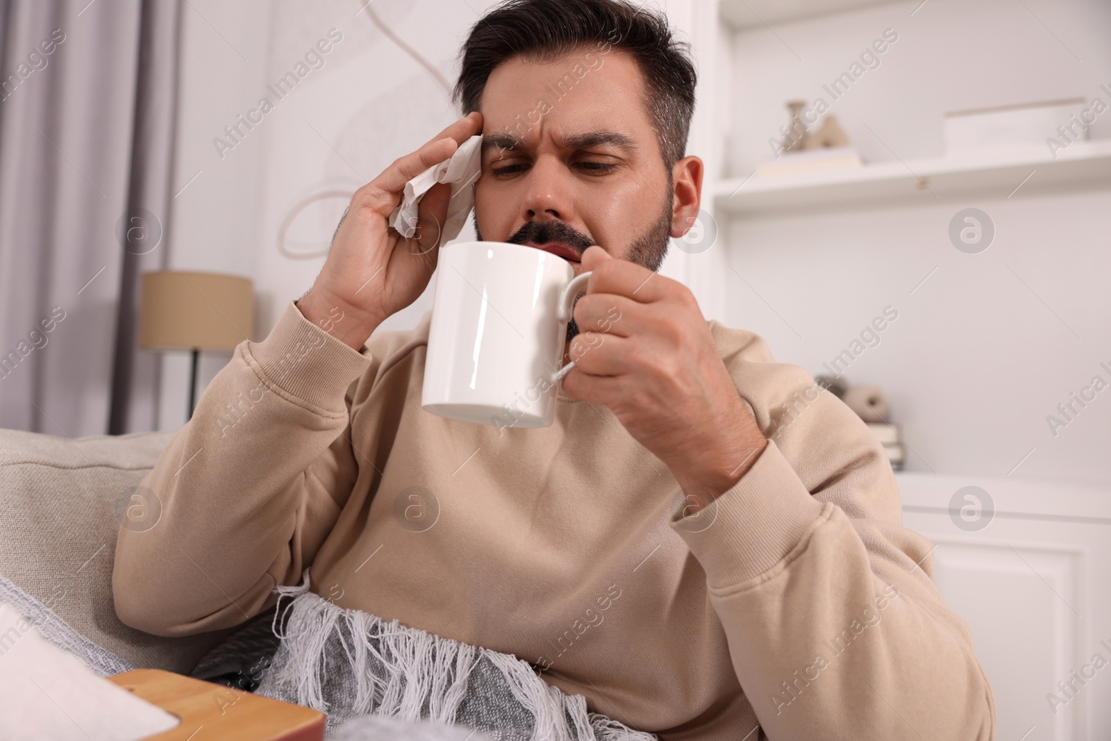 Photo of Sick man with tissue and cup of drink on sofa at home. Cold symptoms