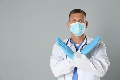 Photo of Doctor in protective mask showing stop gesture on grey background, space for text. Prevent spreading of coronavirus