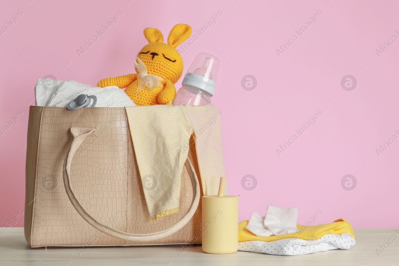 Photo of Mother's bag with baby's stuff on white wooden table against pink background. Space for text