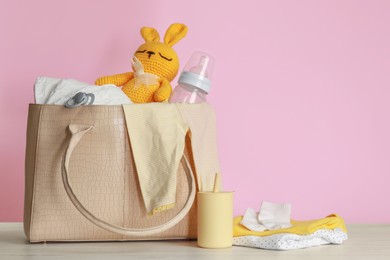 Photo of Mother's bag with baby's stuff on white wooden table against pink background. Space for text