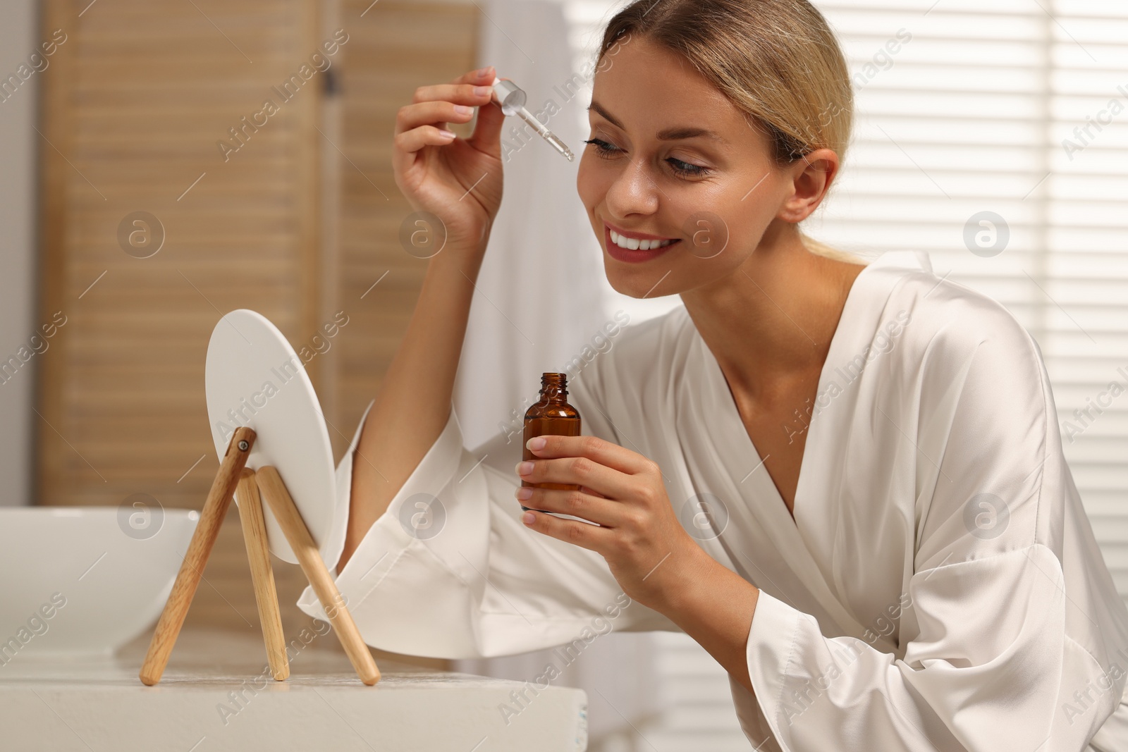 Photo of Beautiful woman applying cosmetic serum onto her face in front of mirror at table in bathroom