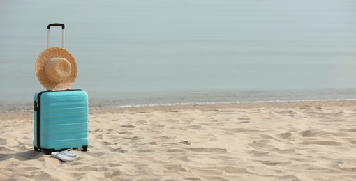 Turquoise suitcase, flip flops and straw hat on sandy beach, space for text