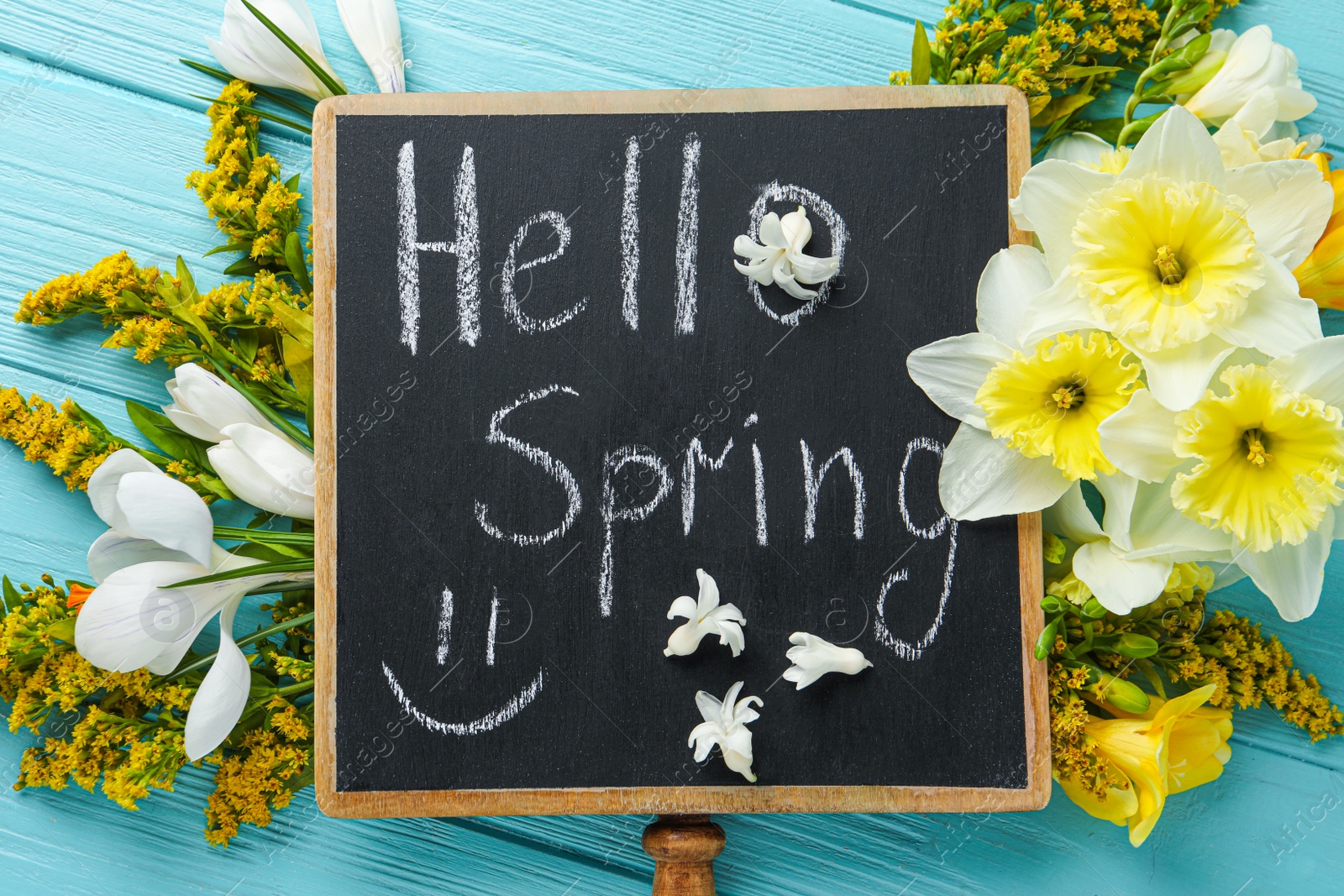 Photo of Blackboard with words HELLO SPRING and flowers on light blue wooden table, flat lay