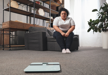Photo of Depressed overweight woman looking at scales in living room