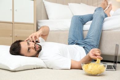 Photo of Lazy young man with laptop, smartphone and bowl of chips lying on floor at home