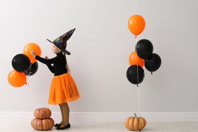 Photo of Cute little girl with balloons and pumpkins wearing Halloween costume near light wall