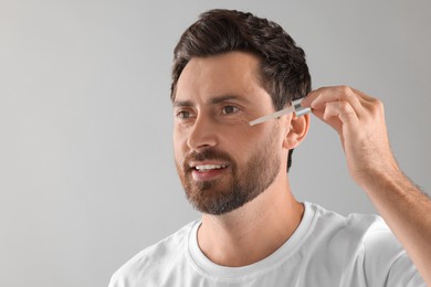 Smiling man applying cosmetic serum onto his face on light grey background. Space for text