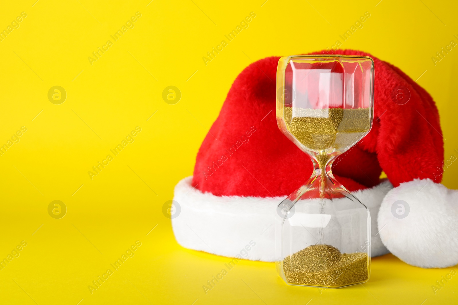 Photo of Hourglass and Santa hat on yellow background, space for text. Christmas countdown