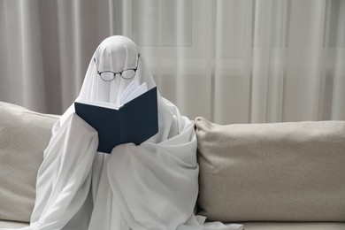 Photo of Creepy ghost. Person covered with white sheet reading book on sofa at home, space for text